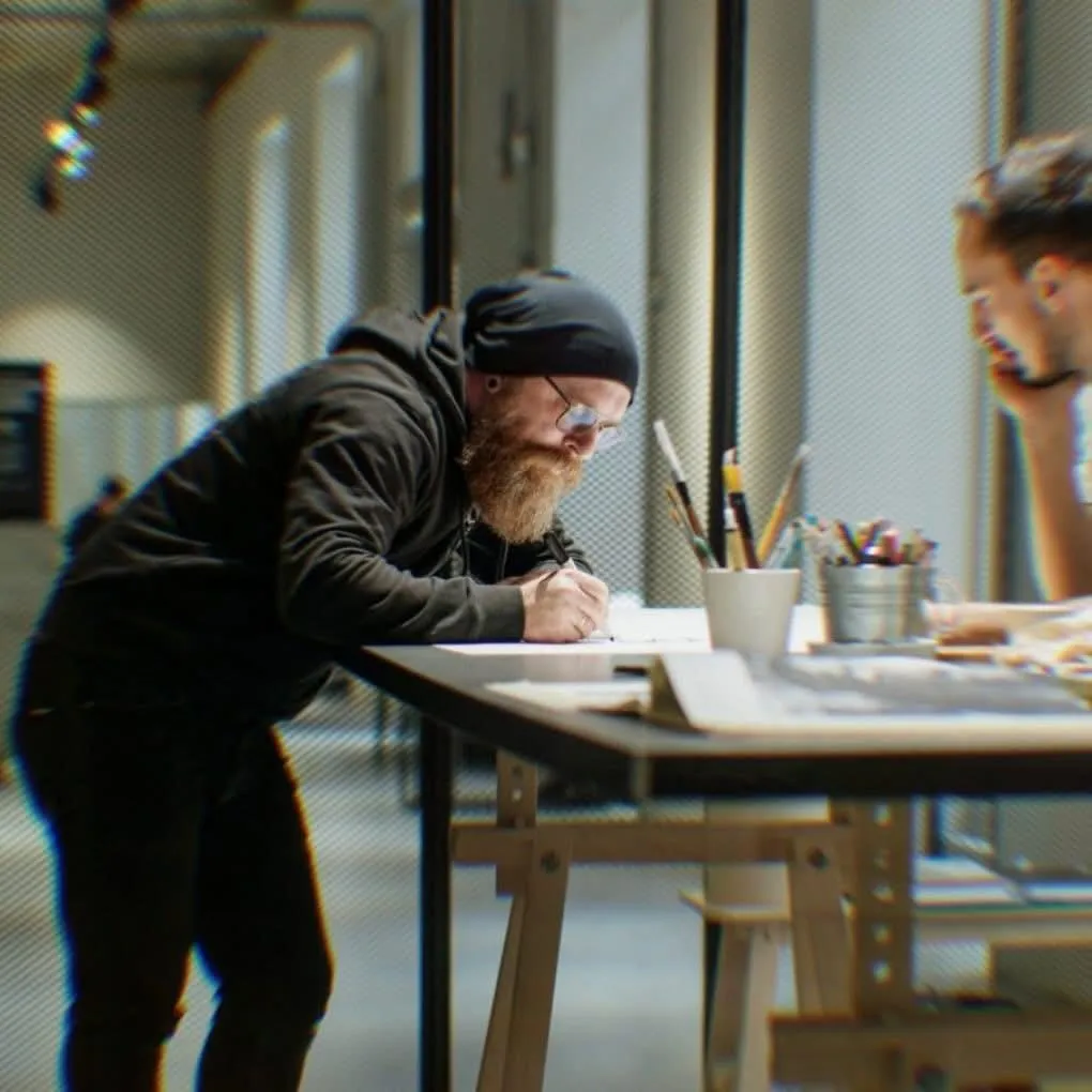 Boti in a black beanie leaning over a long table, drawing with a pen, brushes in a cup beside him