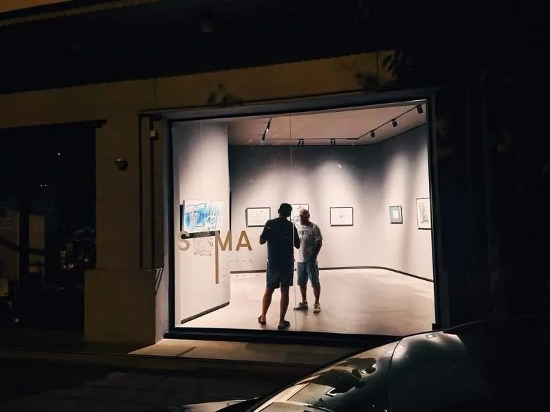 The gallery at night seen through the street window, two visitors inside