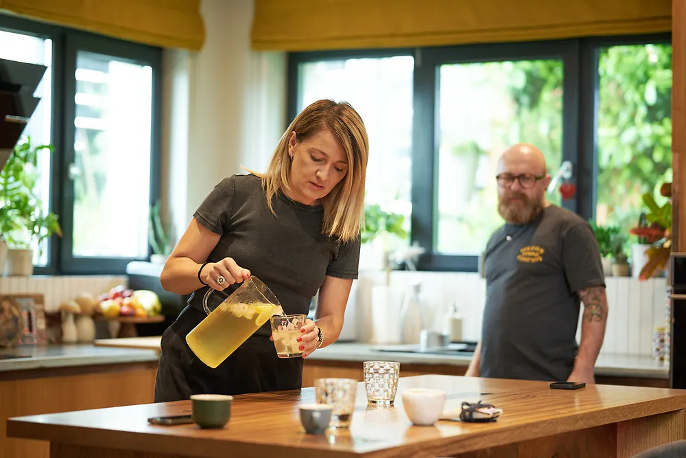 Gyöngyi pouring a drink at the kitchen island while Botond stands by the window