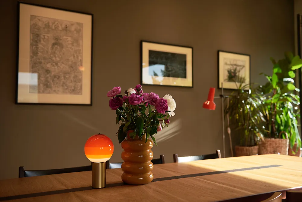 The long dining table with a small orange lamp and a vase of peonies in front of framed prints