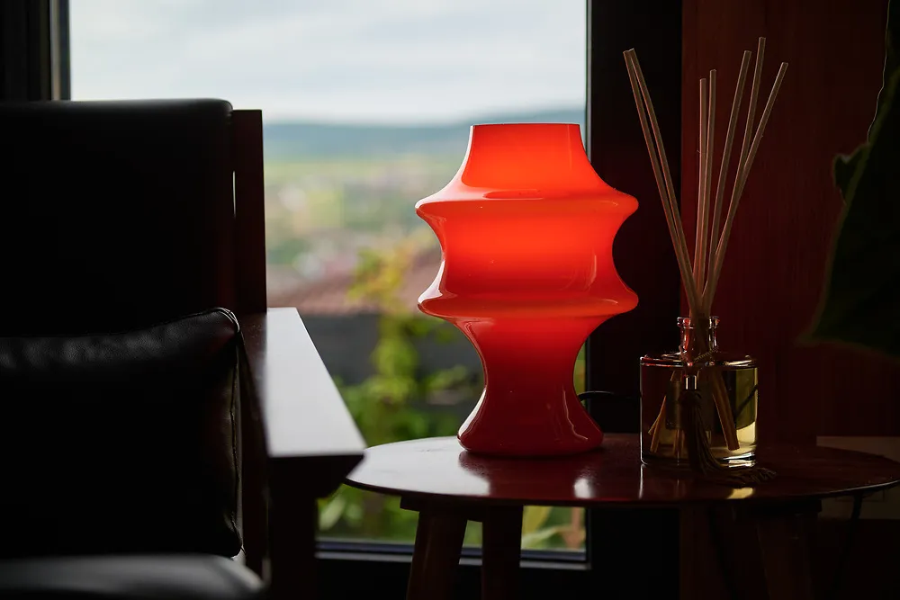 A sculptural red glass lamp on a side table by a window overlooking hills